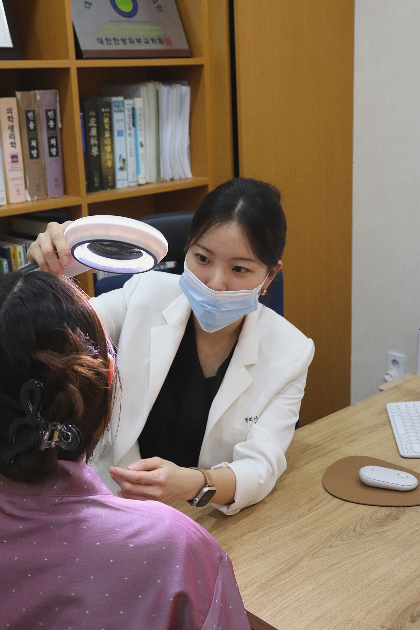 진료실에서 원장이 조명 확대경으로 환자 피부를 살펴보는 상담 장면